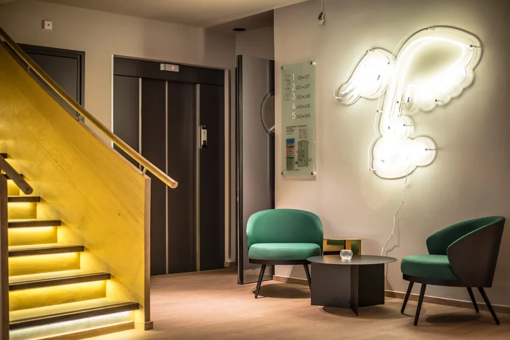 Espace du service Informations utiles de l’Hôtel Rohan Strasbourg avec escaliers éclairés, fauteuils verts et décoration lumineuse murale